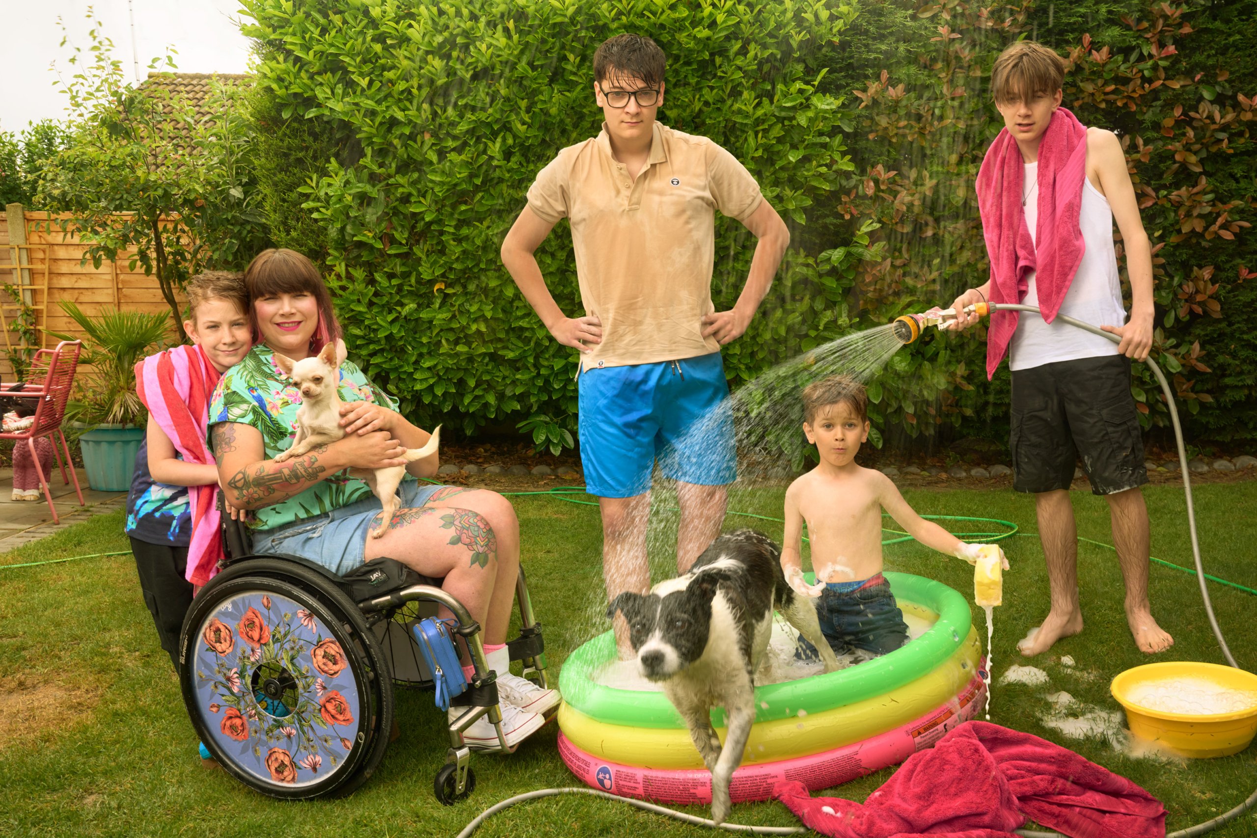 Nina in her garden surrounded by her four kids and their dogs, one of the boy's is filling an inflatable splash pool with water.