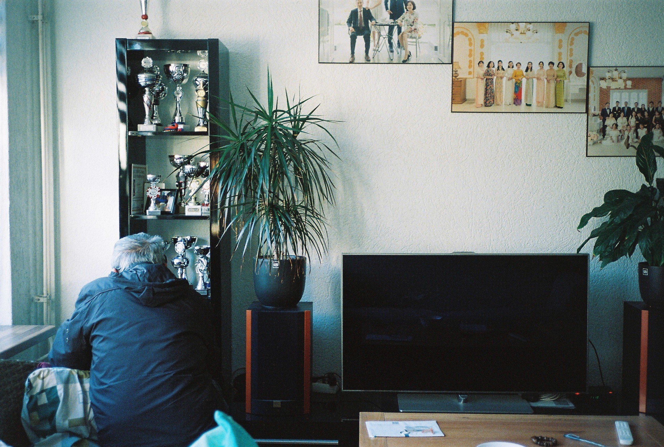 Trams father is sitting in front of a cabinet filled with trophies in their livingroom.