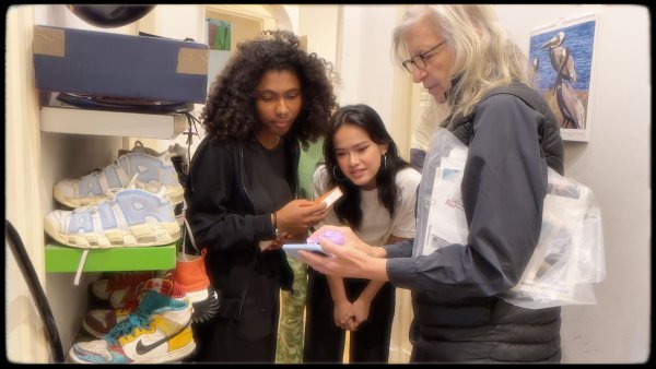 Annie showing something on her phone to Maria and her friend standing in a hallway with shoes on shelves.