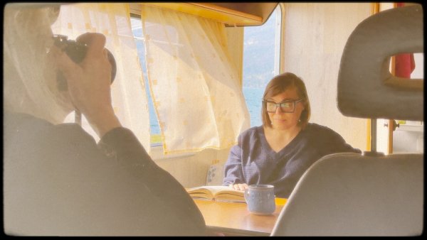 Annie taking photos of Sharon sitting by her dining table reading a book.