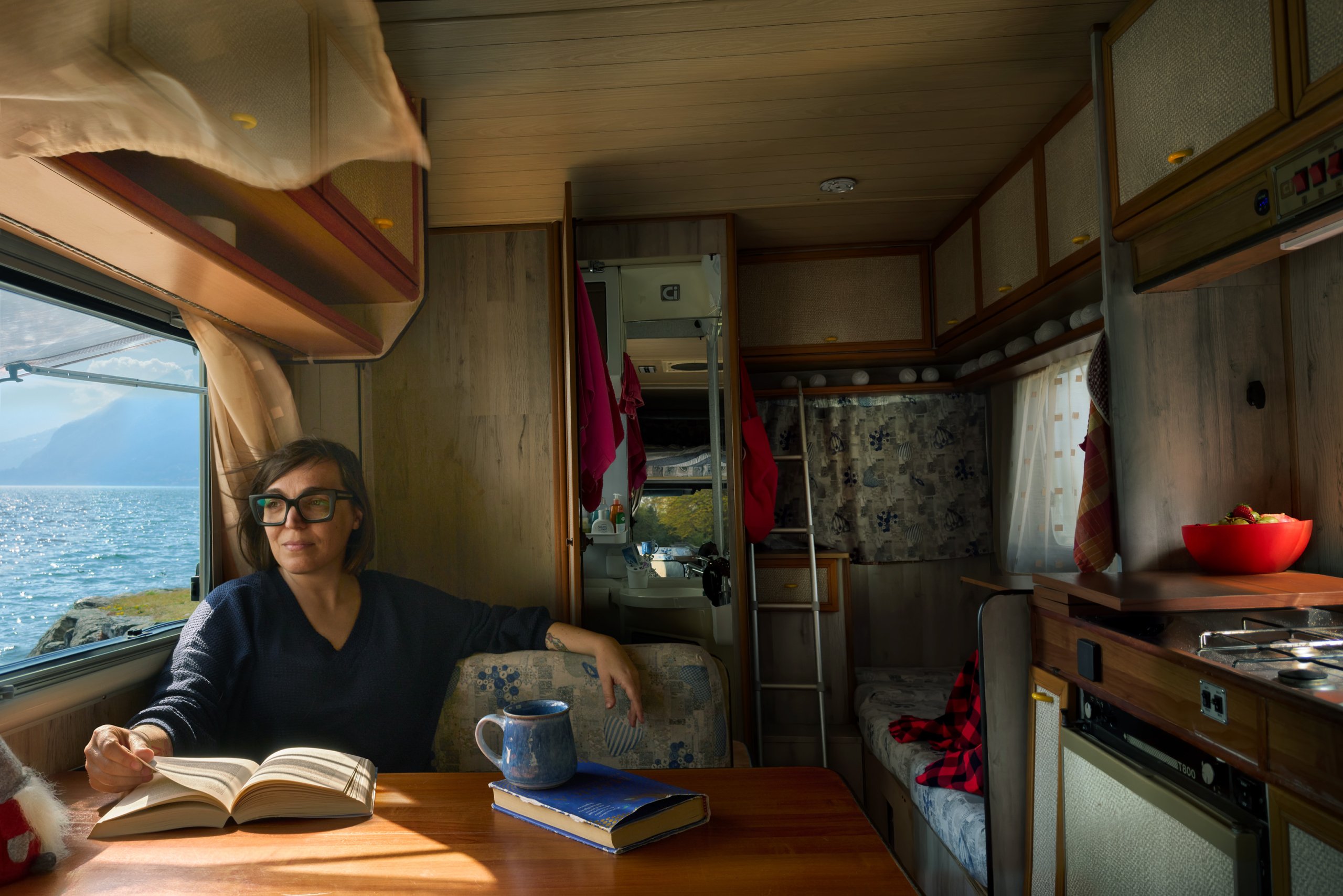Sharon sitting by the table in her camper looking out at the ocean through the window.