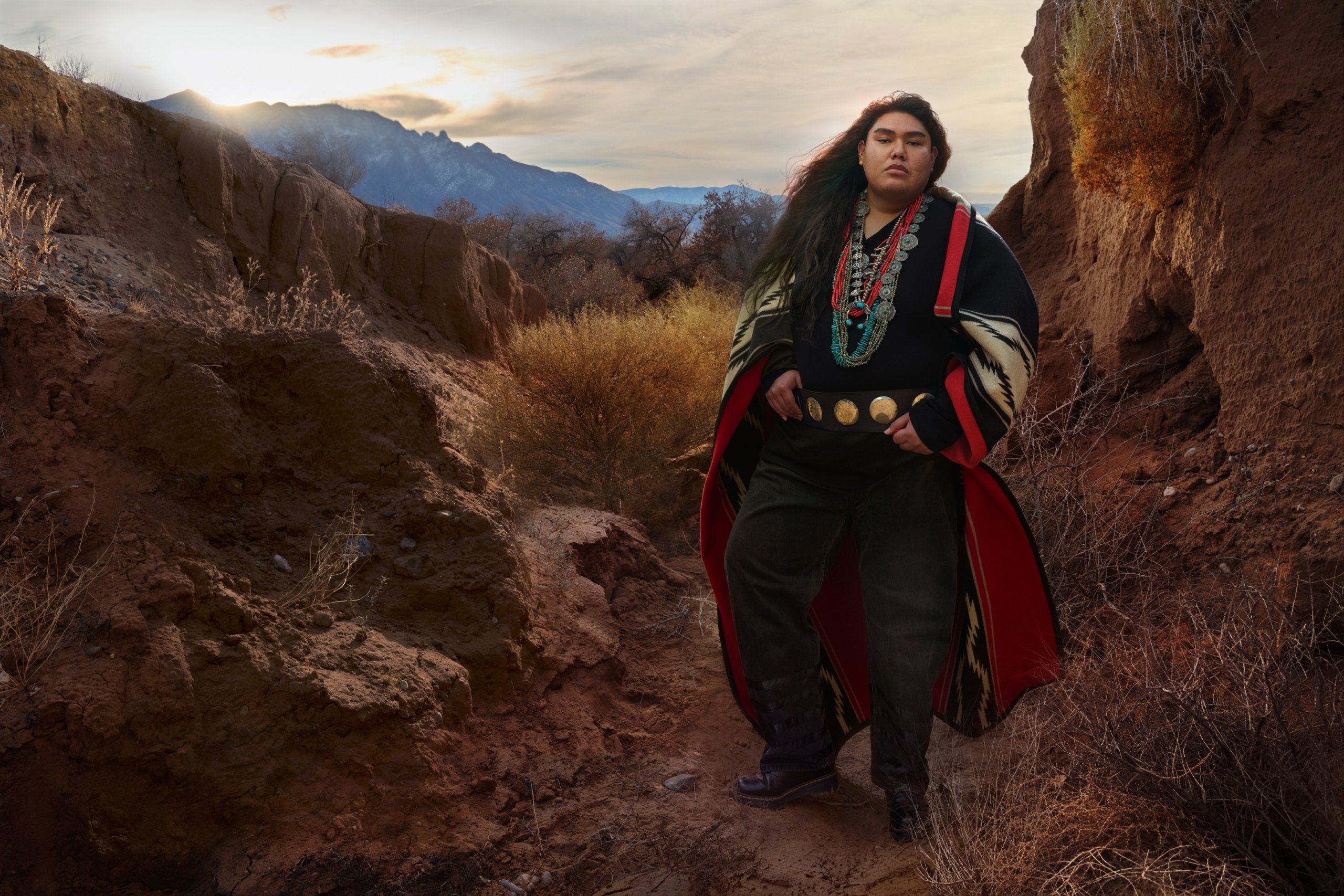 Kymon is standing outside wearing traditional Navajo clothes, large mountains in the background with the sun setting.