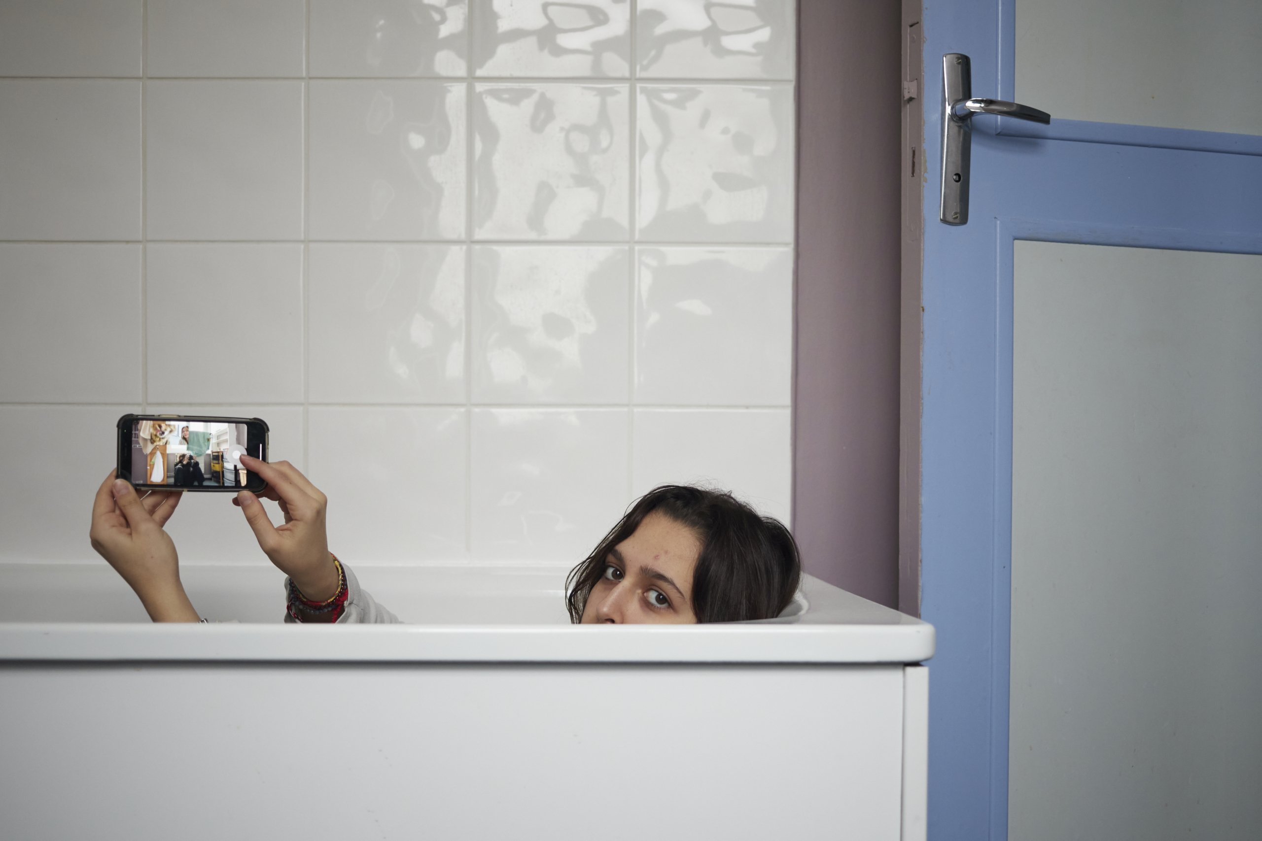 A woman laying in an empty bathtub, shows her phone screen to the photographer.